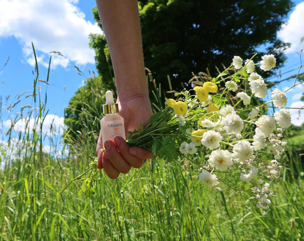 A hand holding VRéserve Bee Glow cosmetic dropper bottle with a gold cap, positioned in front of a bouquet of white and yellow wildflowers, outdoors in a grassy field under a blue sky.