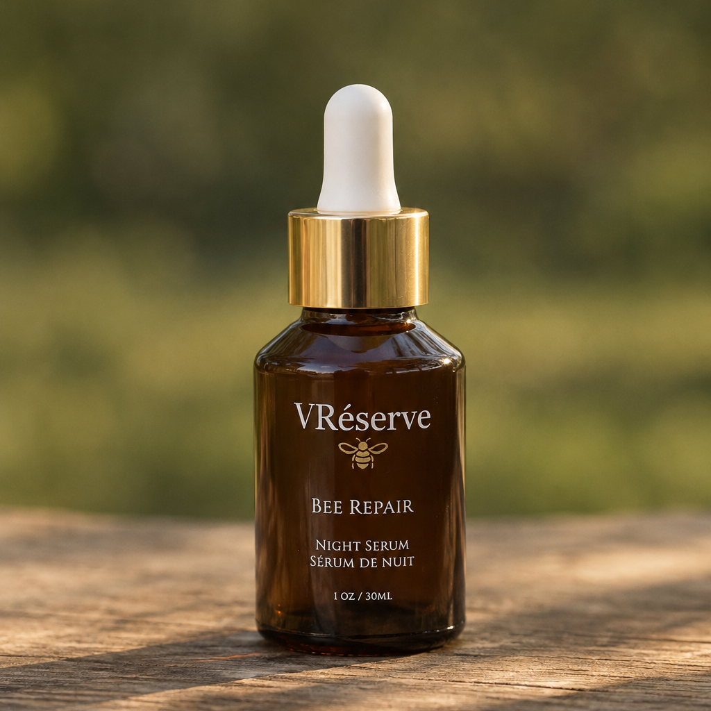 VReserve Bee Repair Night Serum in an amber glass dropper bottle with a gold collar and white dropper top, set on a wooden surface with a blurred green outdoor background.