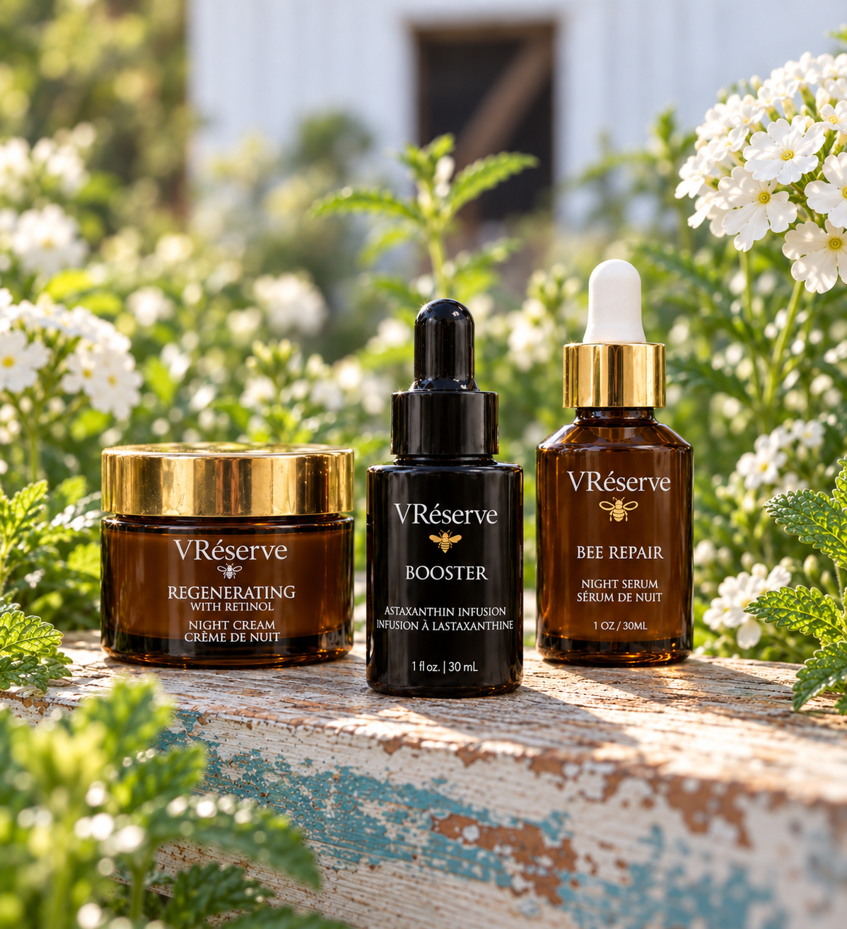 Three VReserve skincare products are arranged on a weathered wooden surface outdoors with green foliage and white flowers in the background. The set includes a Regenerating Night Cream jar with a gold lid, a AstaxanthinBooster dropper bottle in black, and a Bee Repair Night Serum dropper bottle in amber brown with a gold cap and white dropper top.