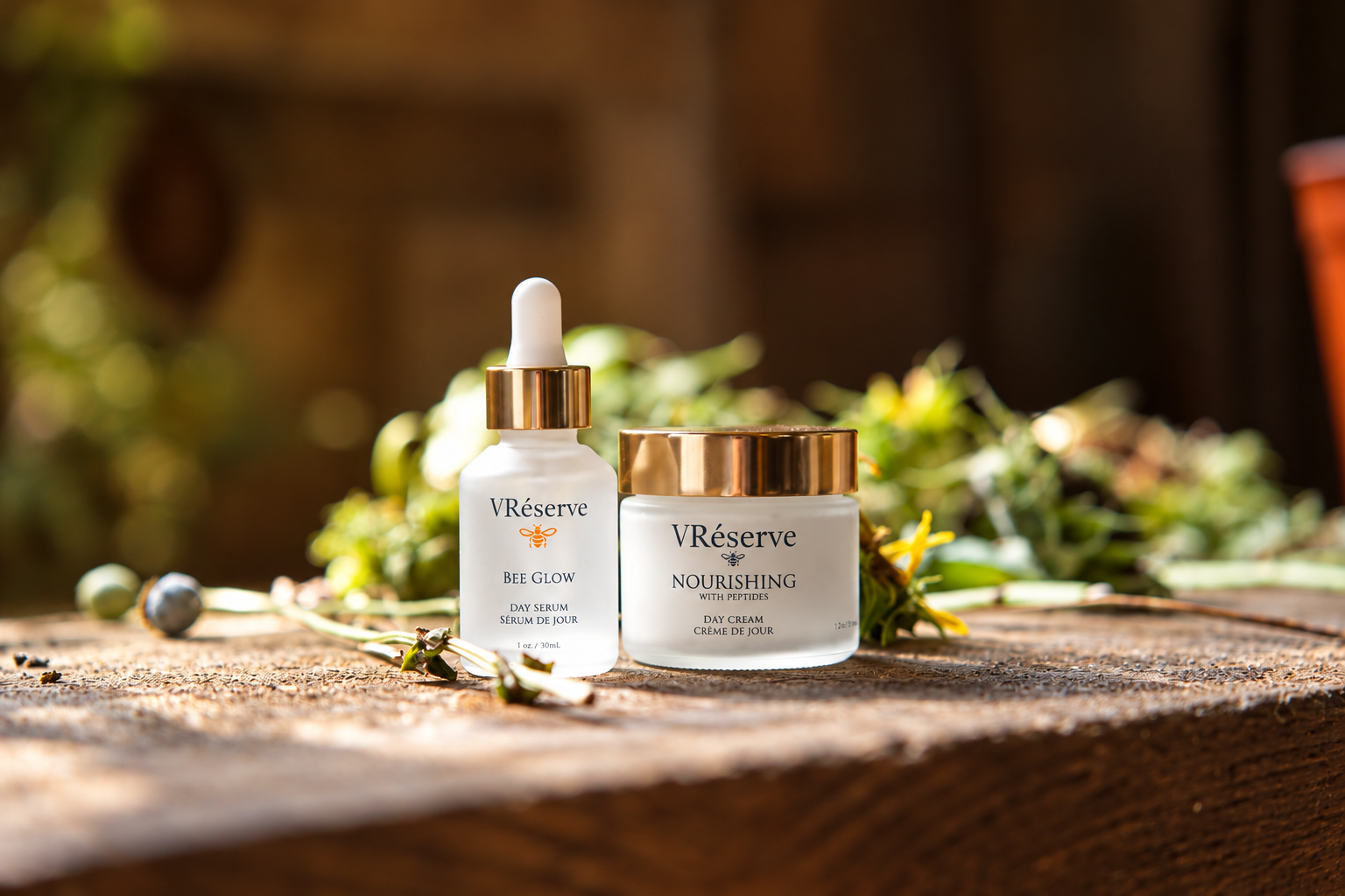 V Réserve skincare set featuring a 'Bee Glow' serum dropper bottle and a 'Nourishing' cream jar, both with white glass and gold lids, displayed on a rustic wooden surface with green leaves and wildflowers.