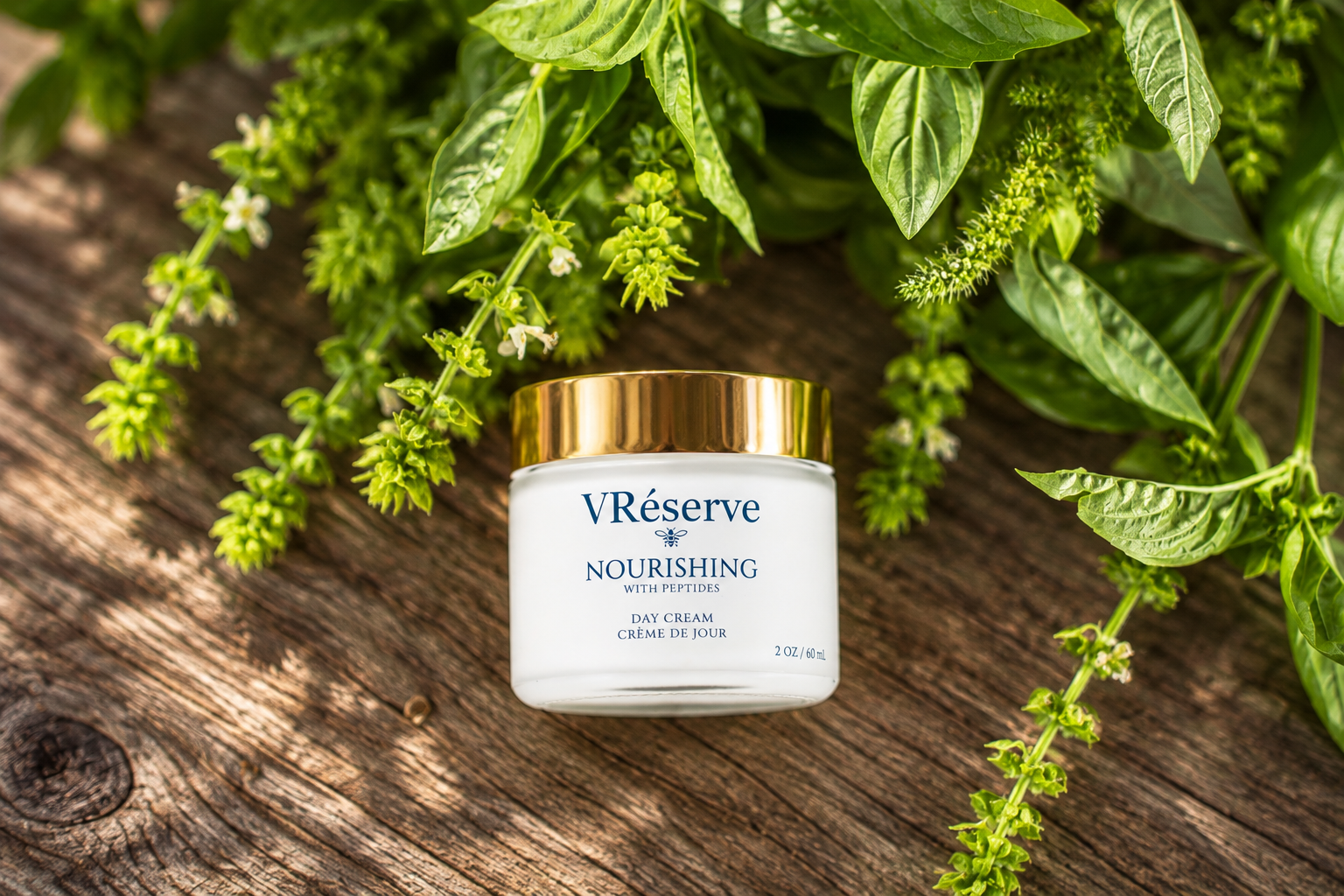A jar of VRésence Nourishing Day Cream sits on a rustic wooden surface surrounded by fresh green basil leaves and small white flowers. The jar is white with a shiny gold lid and blue branding text.