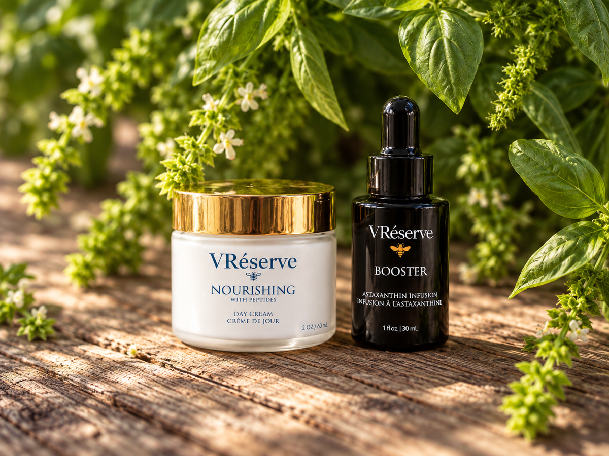 Two skincare products from VRéserve displayed on a rustic wooden surface surrounded by fresh green herbs and small white flowers. The product on the left is a white cream jar with a gold lid labeled 'Nourishing', and the product on the right is a black dropper bottle labeled 'Booster'.