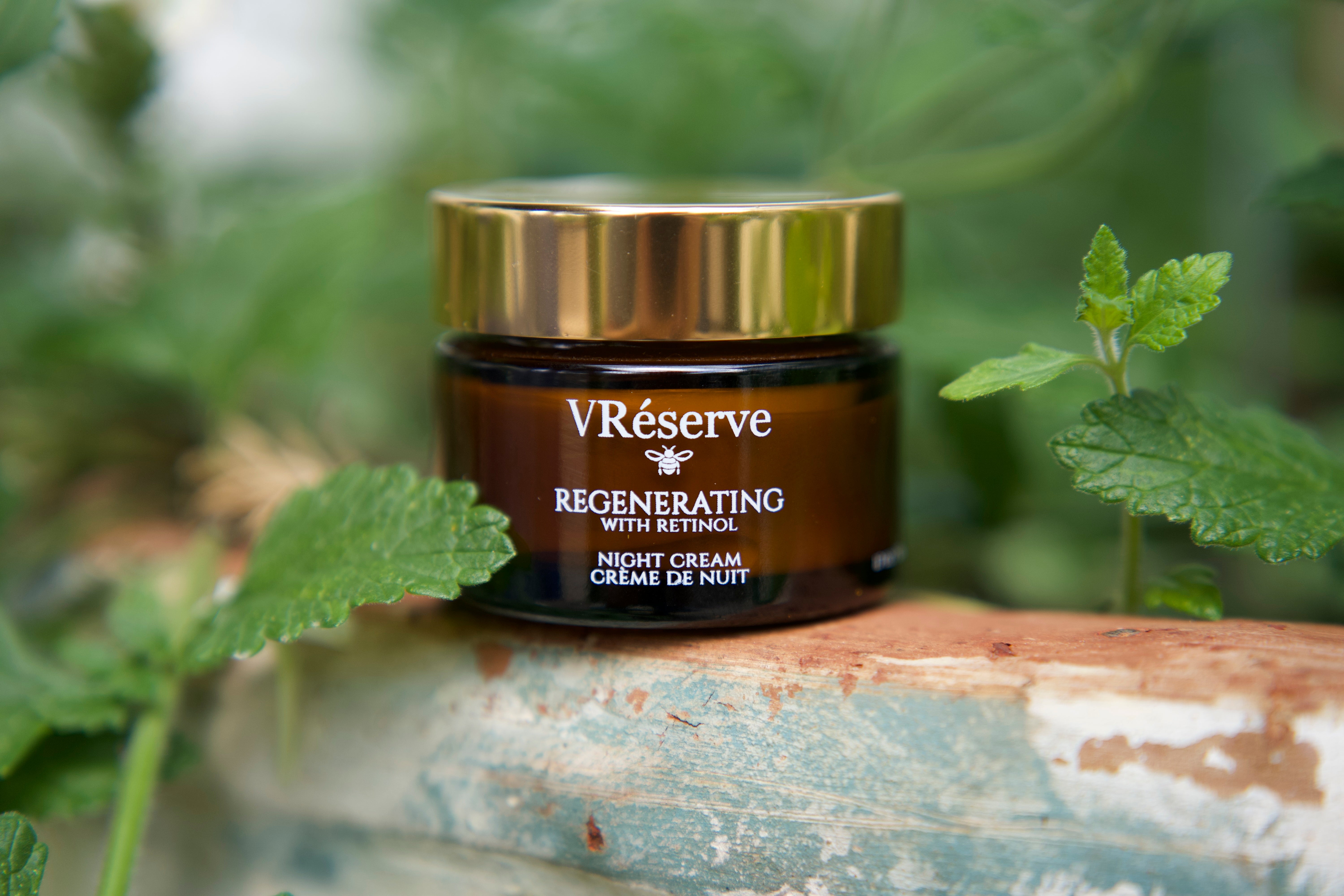 VReserve Regenerating Night Cream in a dark amber jar with a shiny gold lid, set on a rustic wooden surface surrounded by green leaves.