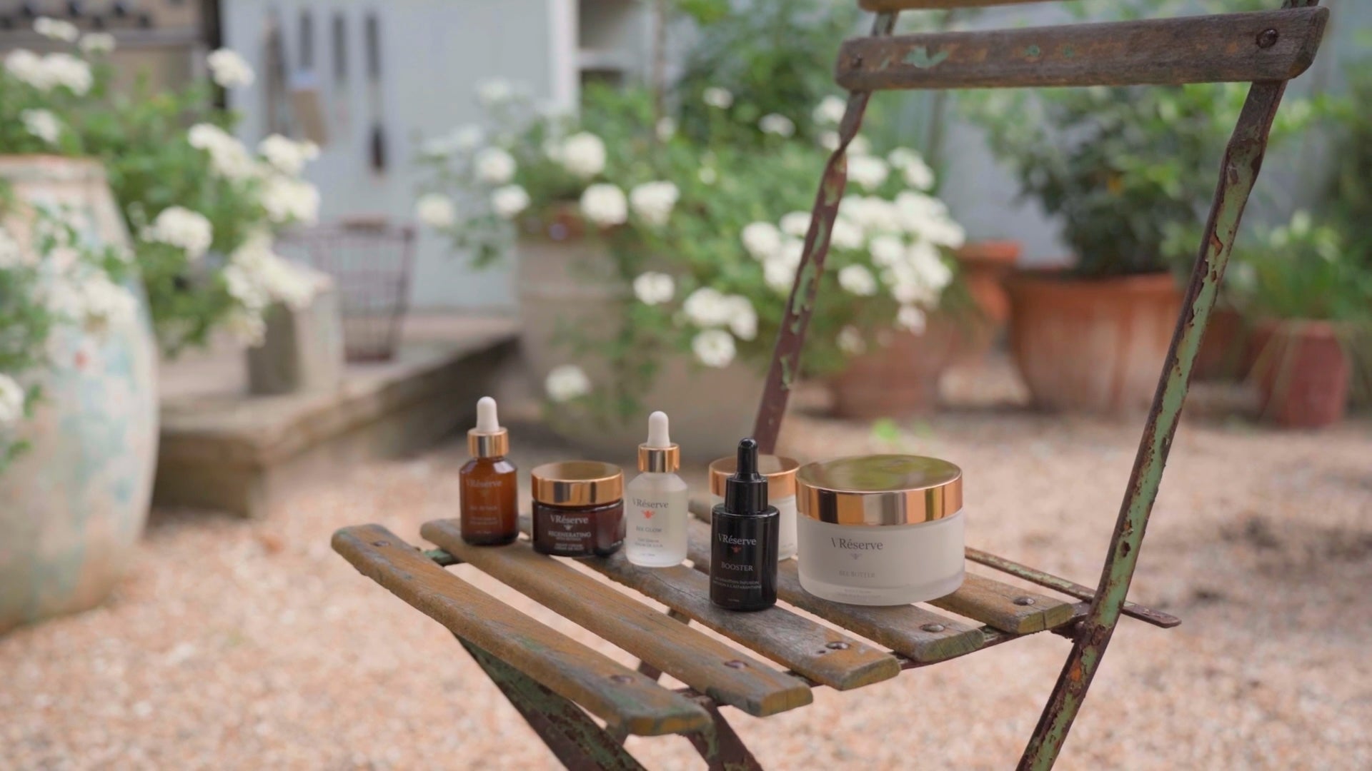 Entire collection of VReserve skincare products, including dropper bottles and a cream jar, arranged on the slats of a rustic wooden garden chair in an outdoor patio setting.
