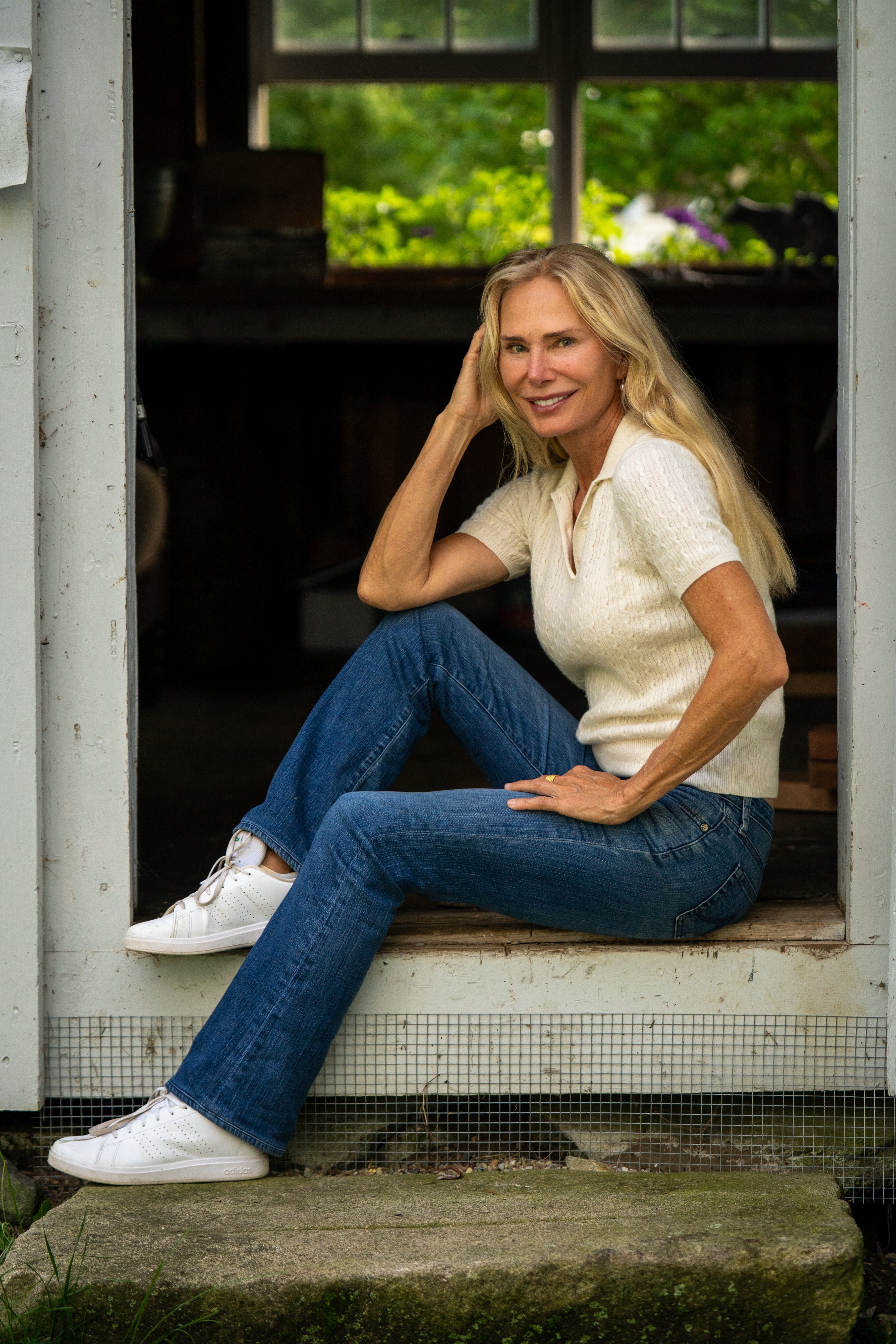 Valesca Guerrand-Hermes sits on the threshold of a rustic building, wearing a cream short-sleeve knit top, blue jeans, and white sneakers.