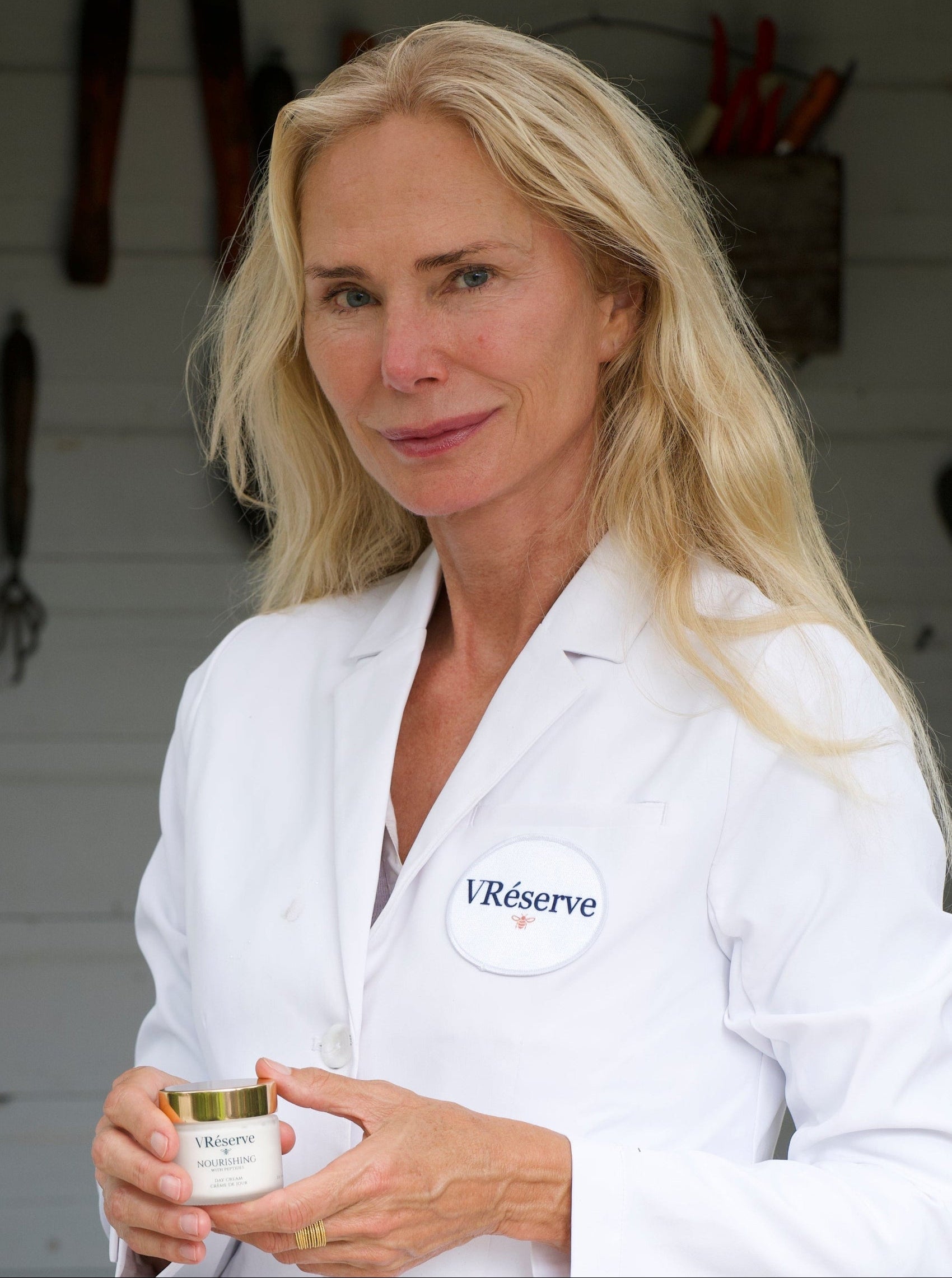Valesca Guerrand Hermes wearing a white lab coat with VRéserve logo badge stands in front of a white shed background, holding a VRéserve Nourishing Day Cream.