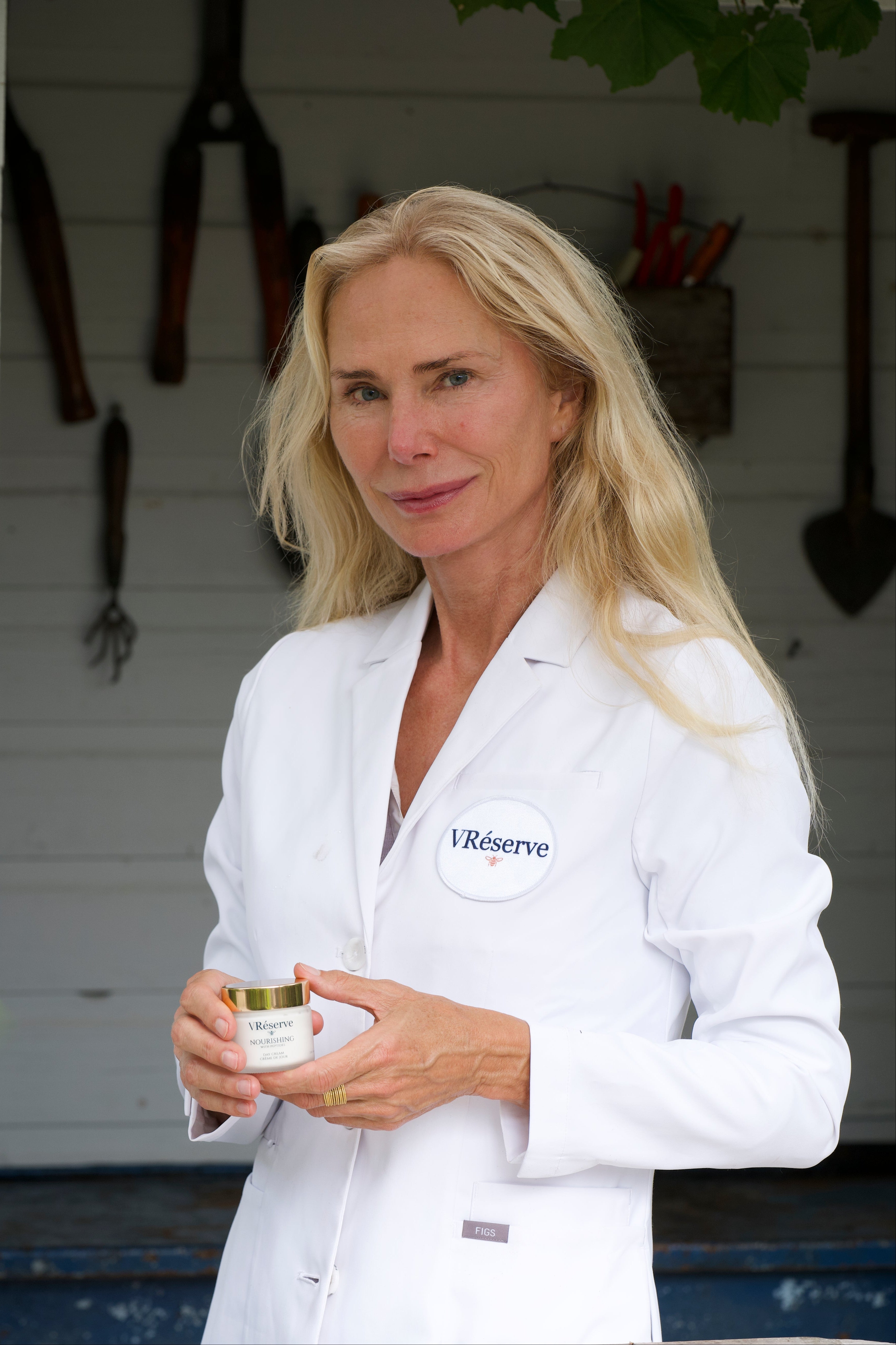 A blonde woman, Valesca Guerrand Hermes wearing a white lab coat with a 'VRESERVE' logo badge stands in front of a white shed background, holding a vreserve day cream.