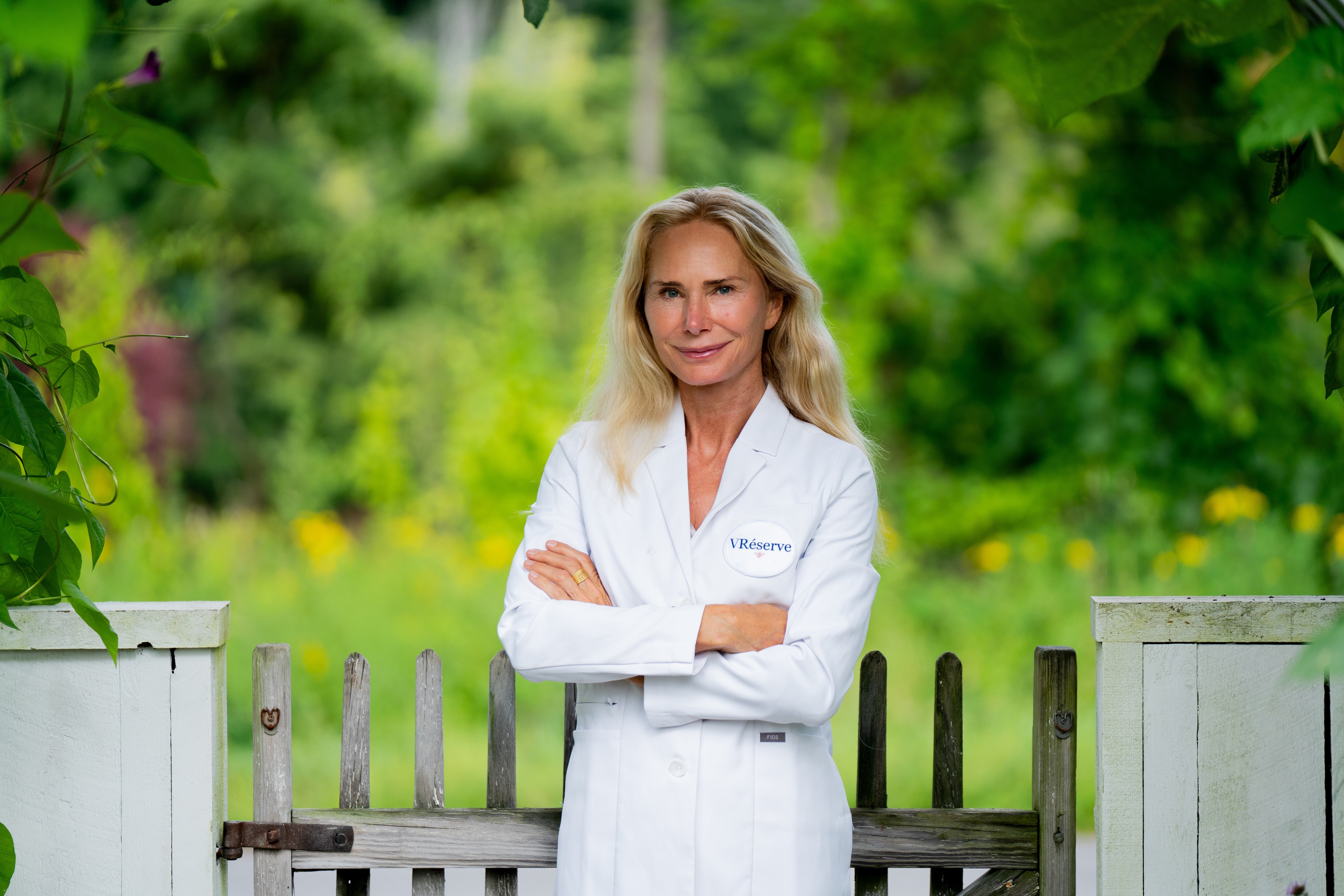 Valesca Guerrand Hermes in a white medical lab coat stands with arms crossed in front of a white picket fence, with a lush green garden background.