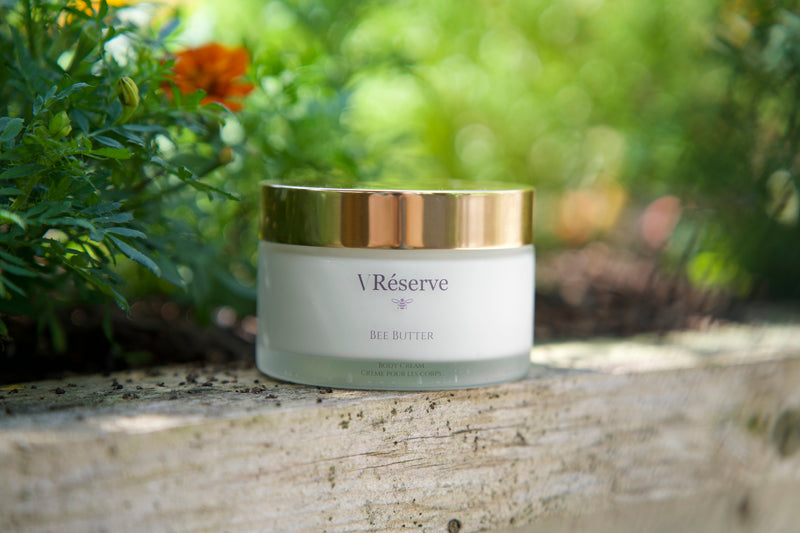 A jar of Vreserve Bee Butter with a white body and a metallic gold lid, resting on a weathered wooden surface with a blurred garden background.