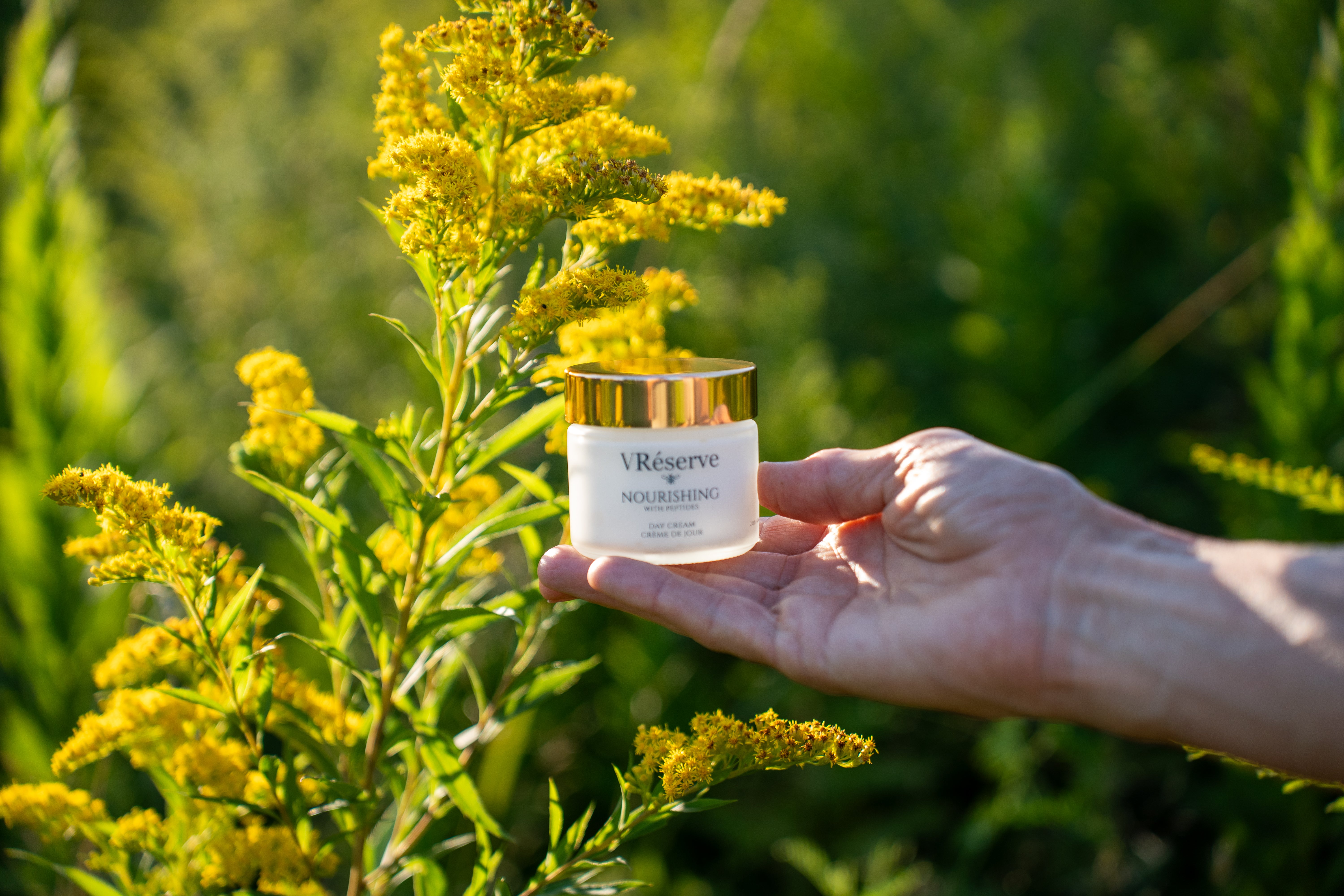 A hand holding a small white cosmetic jar with a gold lid labeled VRéserve Nourishing, set against a blurred outdoor background of green foliage and yellow flowers.