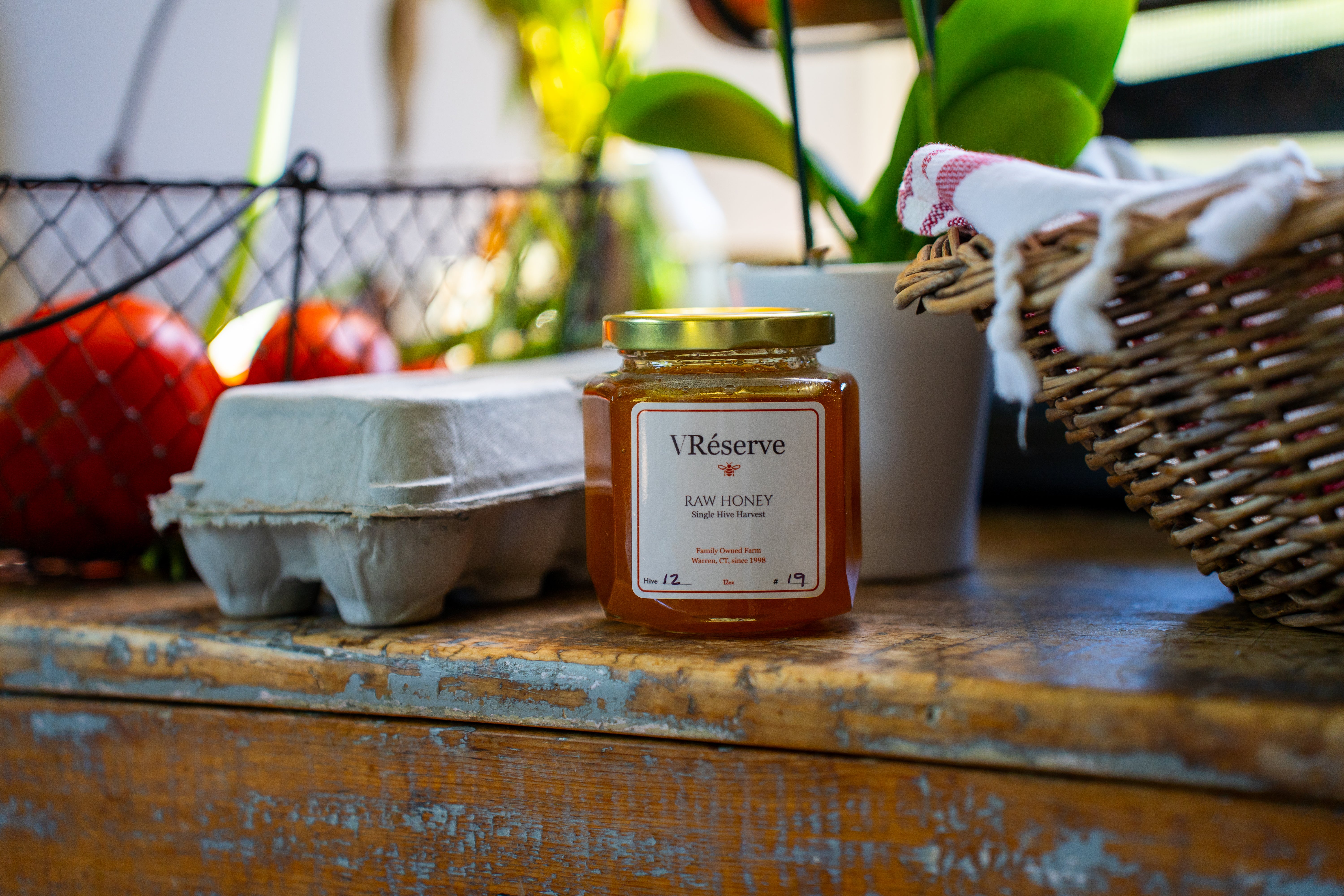 A jar of VRéserve Raw Honey placed on a rustic wooden table, surrounded by farm-style props including a carton, tomatoes, and a wicker basket.
