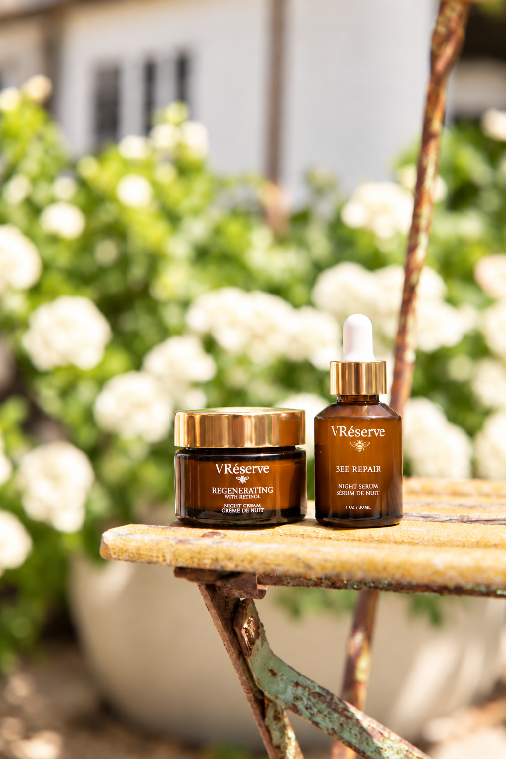 Two VRéserve skincare products, a Regenerating Night Cream jar and a Bee Repair serum dropper bottle, displayed on a rustic wooden table with a blurred garden background of white flowers.