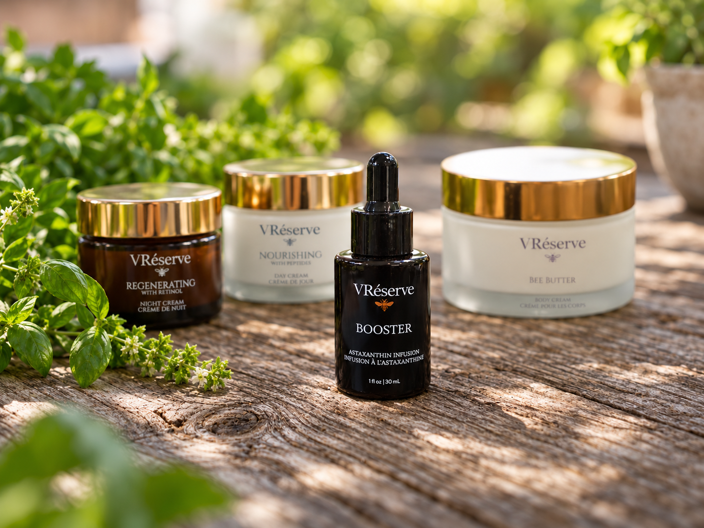 Four VRéserve skincare products arranged on a rustic wooden surface with fresh green herbs in the foreground and a softly blurred outdoor garden background. The products include a black dropper bottle labeled BOOSTER in front, with Night Cream, Bee Butter, and Day Cream, jars behind it.