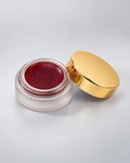 VReserve Bee Balm in a small round cosmetic jar with a translucent frosted base showing a deep red cream product inside, next to its shiny gold lid on a light gray background.