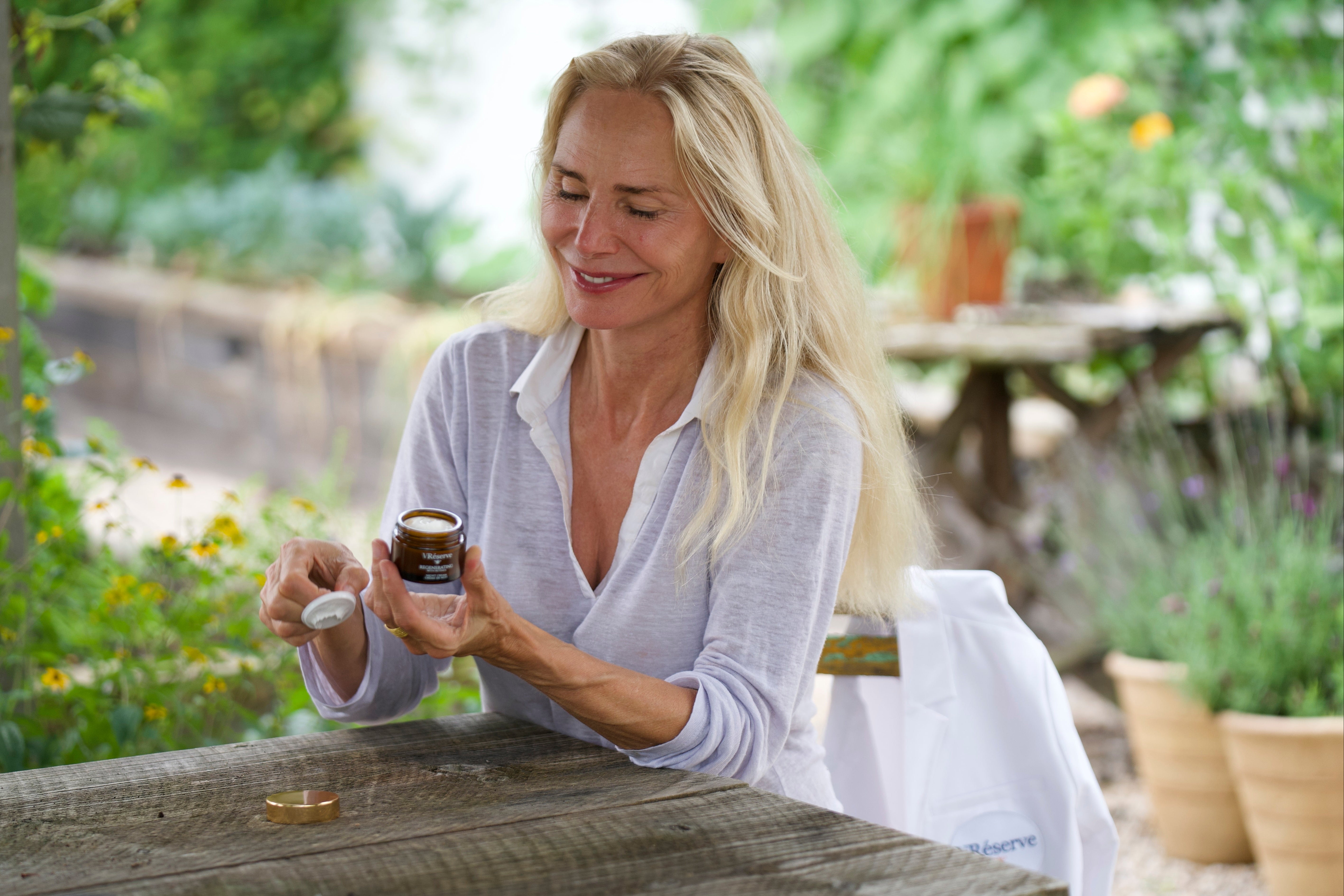 A woman, Valesca Guerrand-Hermes, in a white shirt sits at a wooden table in a garden setting, holding an open vreserve night cream. A white garment bag rests on the table beside her.
