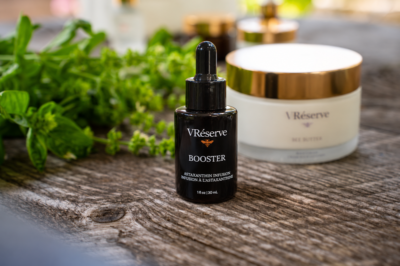 A small black dropper bottle labeled VRéserve Booster stands on a rustic wooden surface, with a cream-colored jar of VRéserve Be Butter behind it and green herbs in the background.