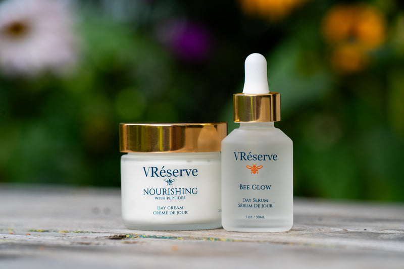 Two VRéserve skincare products on a weathered wooden surface with a blurred garden background: a white jar of Nourishing Day Cream with a gold lid, and a frosted glass dropper bottle of Bee Glow Day Serum with a gold collar and white dropper top.