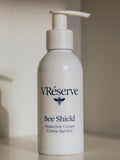 A white pump bottle of VRéserve Bee Shield Protective Cream with a dark navy blue logo and text, set against a soft beige background.