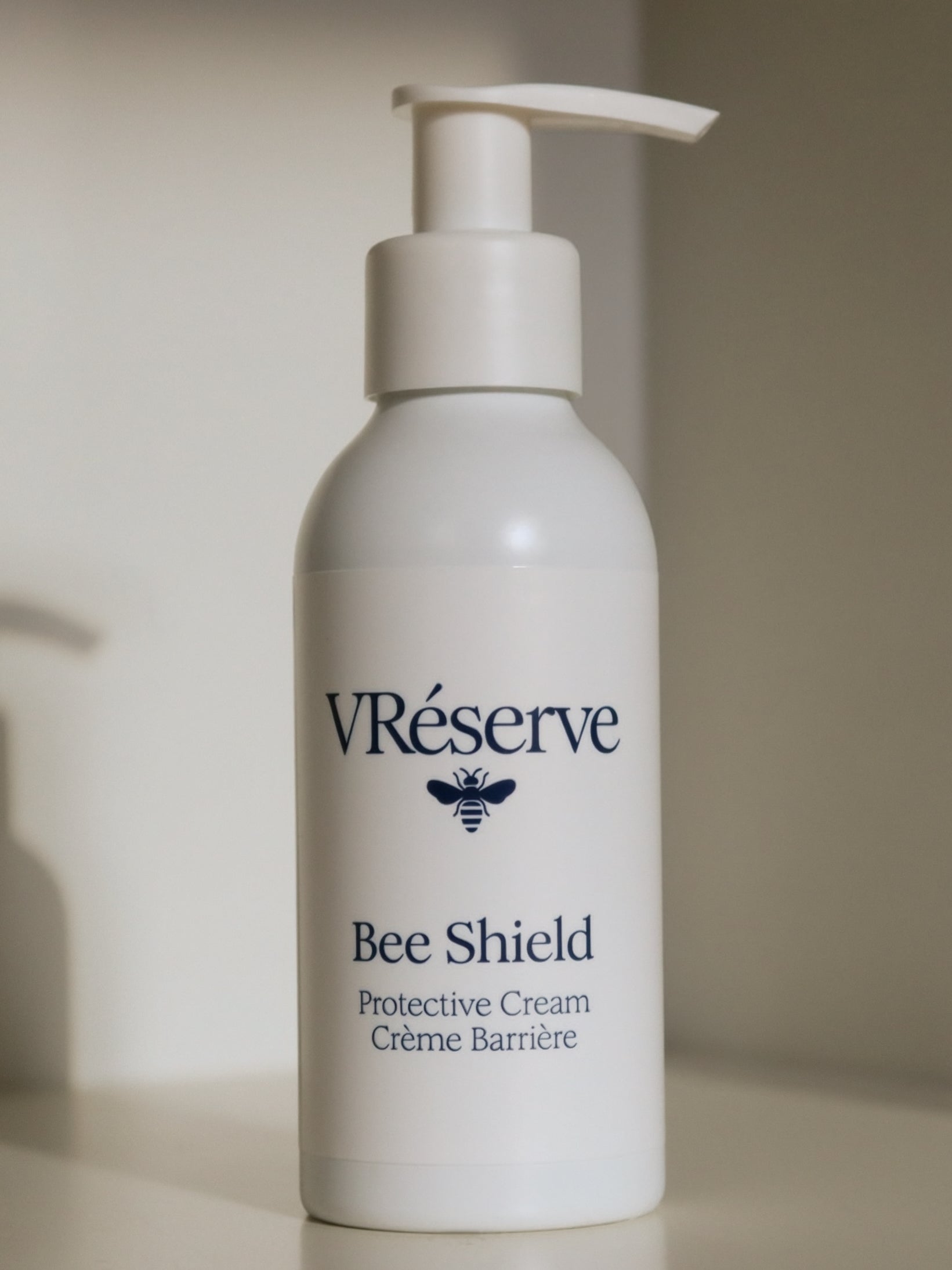 A white pump bottle of VRéserve Bee Shield Protective Cream with a dark navy blue logo and text, set against a soft beige background.
