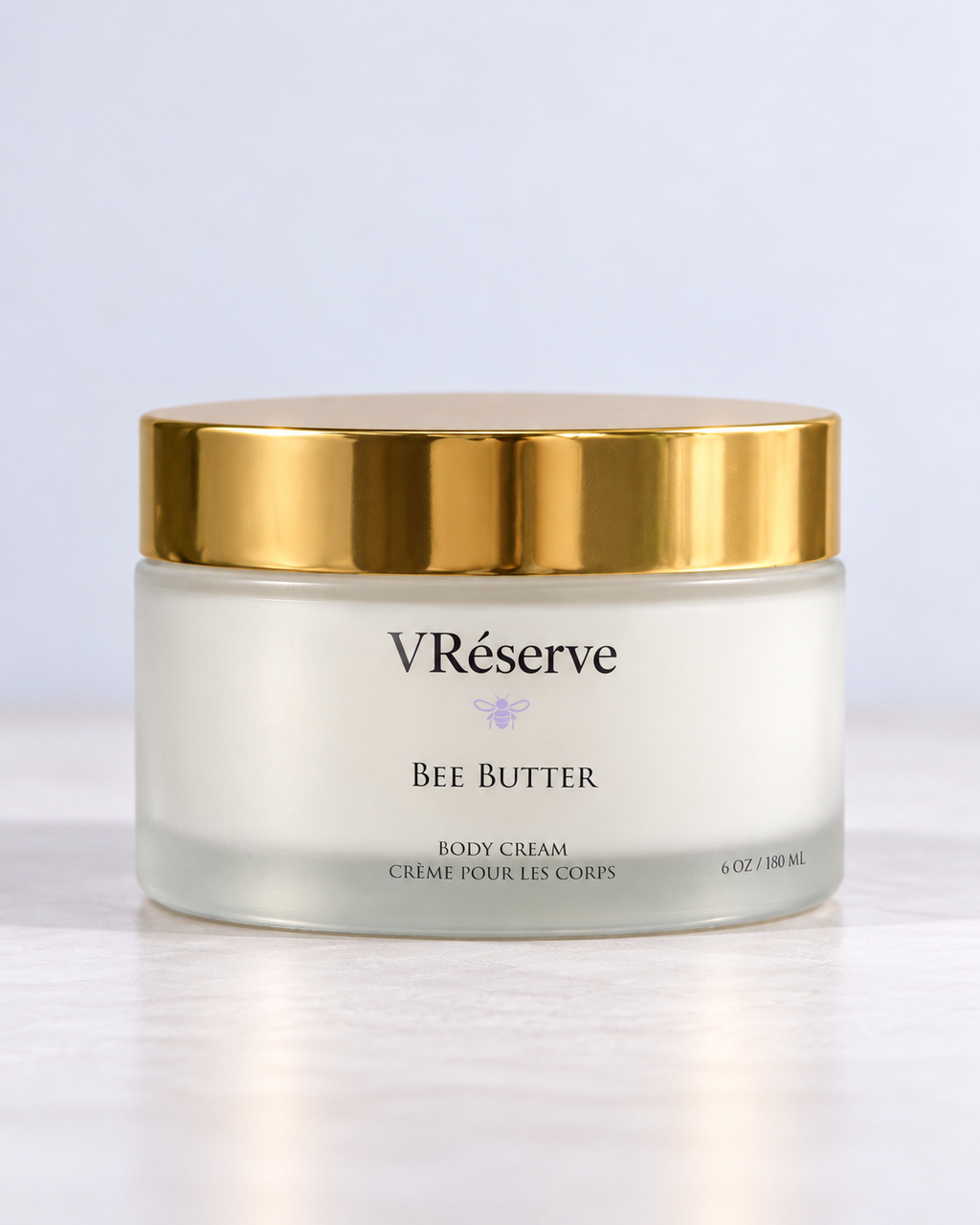 A jar of VRéserve Bee Butter body cream with a white cream inside a frosted glass jar, featuring a shiny gold lid, set against a clean white background.