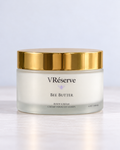 A jar of VRéserve Bee Butter body cream with a white cream inside a frosted glass jar, featuring a shiny gold lid, set against a clean white background.