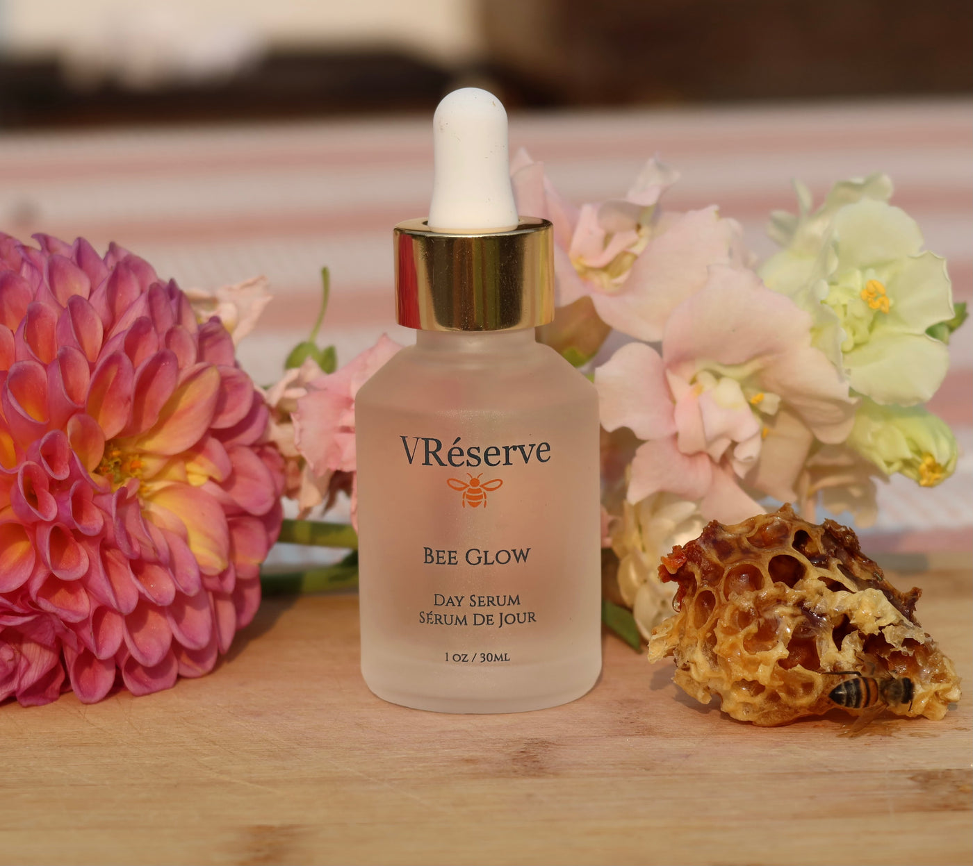 VReserve Bee Glow Day Serum in a frosted glass dropper bottle with a gold collar and white dropper cap, styled on a wooden surface with pink flowers and a honeycomb piece in the background.