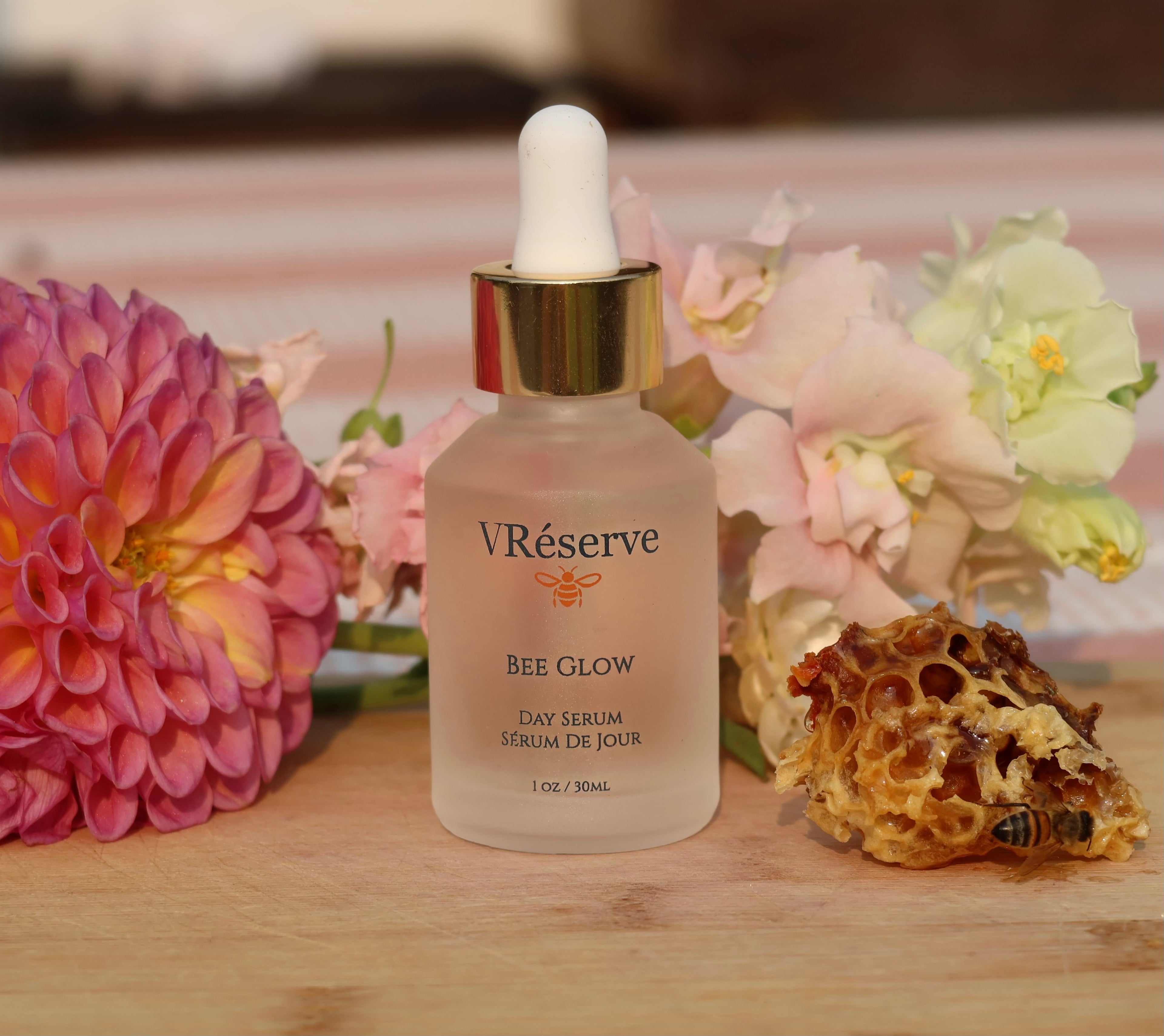 VReserve Bee Glow Day Serum in a frosted glass dropper bottle with a gold collar and white dropper cap, styled on a wooden surface with pink flowers and a honeycomb piece in the background.