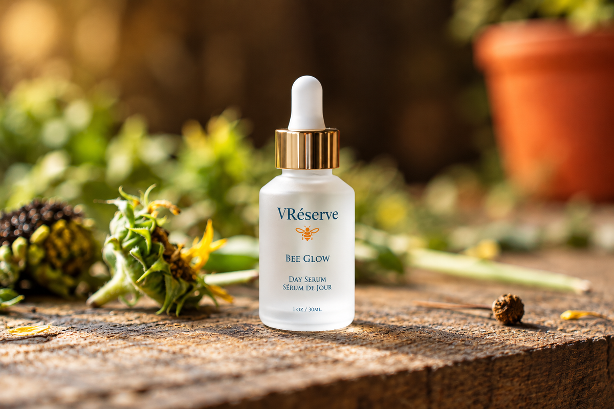 A bottle of VRéserve Bee Glow Day Serum stands on a rustic wooden surface outdoors, surrounded by blurred greenery and dried flowers. The bottle is frosted white with a gold dropper cap and blue branding text.