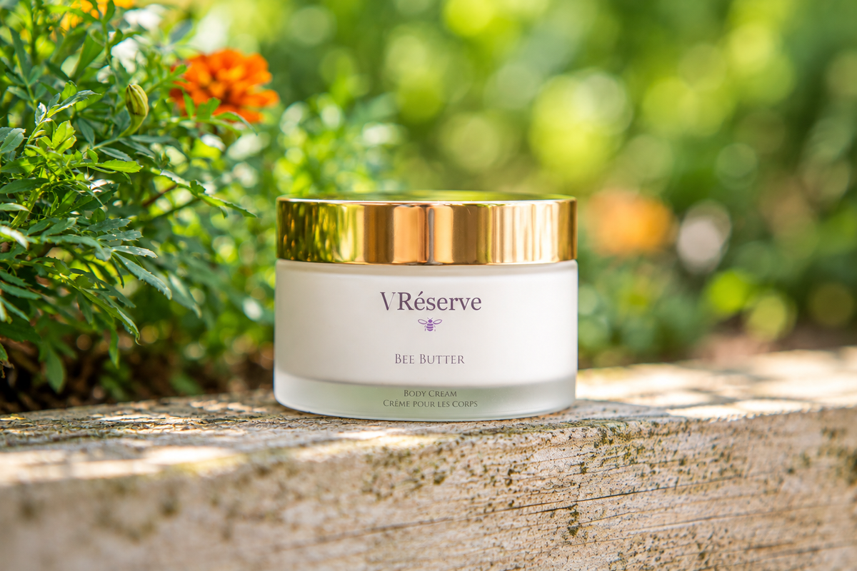 V Réserve Bee Butter cream jar with a gold lid and white body, placed on a weathered wooden surface with green garden foliage and an orange flower in the background.
