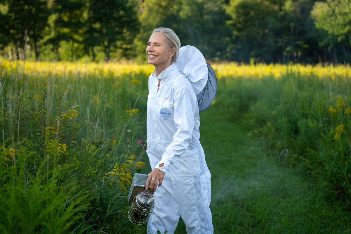 Valesca Guerrand-Hermes in a white beekeeping suit walks through a grassy field holding a smoker and wearing a protective hood. Trees and yellow wildflowers are visible in the background.