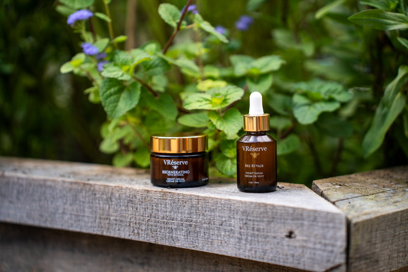 Two amber glass skincare bottles from VReserve, including a jar and a dropper bottle, displayed on a weathered wooden surface with a blurred green garden background.
