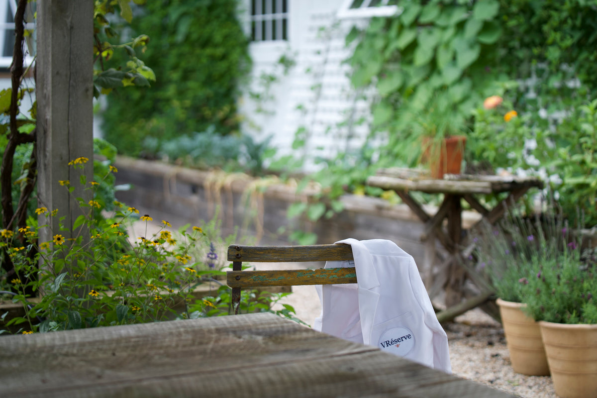 A white lab coat with 'VRESERVE' printed in blue text hanging from a rustic wooden chair in a lush, green garden patio setting.