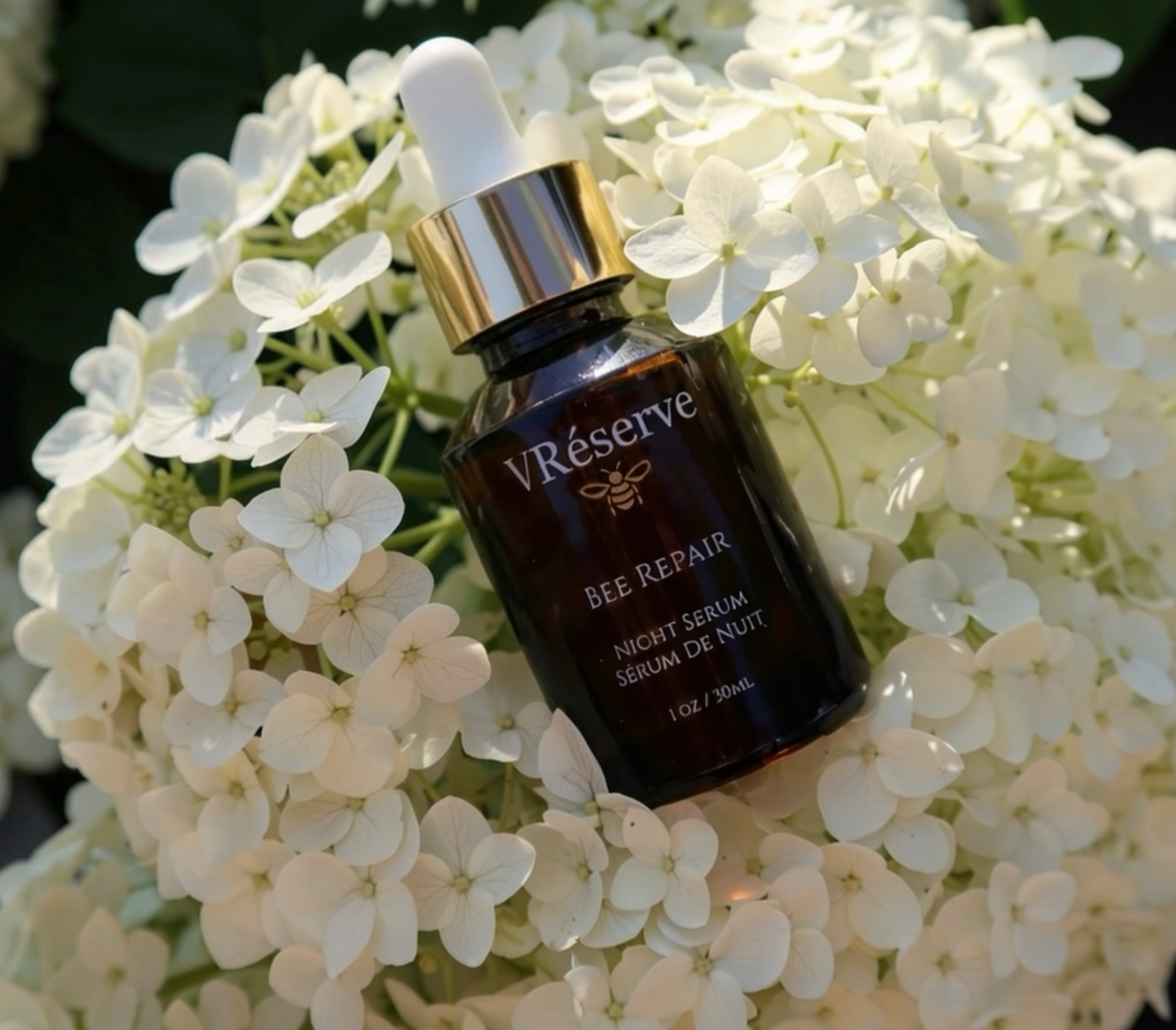 vreserve Bee Repair Night Serum in a dark amber glass dropper bottle with a gold collar, nestled among white hydrangea flowers.